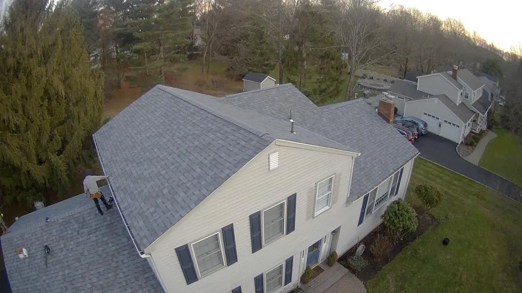 Aerial view of a finished grey architectural shingle roof on a two-story colonial, with a roofer on the lower roof
