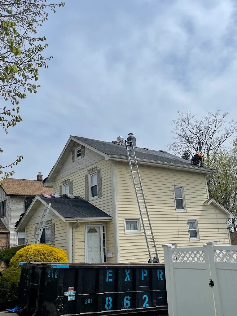 A yellow two-story house with ladders up and a roofer working at the chimney